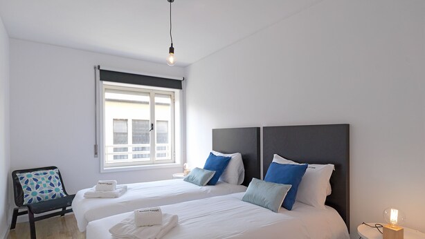 Hotellihuone, jossa on kaksi sänkyä, ikkuna, tuoli ja yöpöytä, jossa on lamppu.