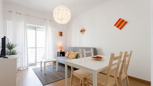 A modern living room with a white sofa, a dining table with wooden chairs, a hanging light fixture, and a small side table with a lamp. - FLH Marquês Bright Apartment (Lisbon)