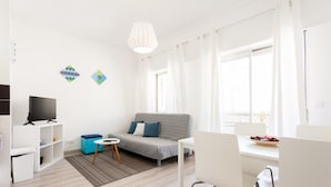 Una sala de estar moderna con un sofá gris, un mueble de televisión blanco y una zona de comedor con una mesa y sillas blancas.