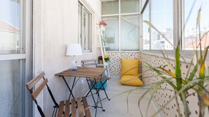 Un balcón con una mesa de madera, sillas, un cojín amarillo y una lámpara blanca.