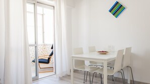 Un comedor moderno con una mesa y sillas blancas, un vidrio que da a un balcón con una silla azul, y una obra de arte geométrica en la pared.