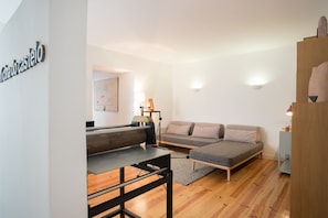 A modern, minimalist room with a wooden floor, a grey sofa, a small bed, a desk with a lamp, and a wall-mounted light fixture.