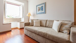 A living room with a beige sofa, a window with a view, and a framed picture on the wall. - FLH Olivais Cozy Flat (Lisbon)