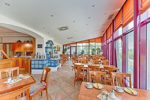 Un vaste espace dédié à la salle à manger, avec de nombreuses tables et chaises en bois, un bar central doté d’un comptoir bleu et de grandes fenêtres laissant entrer la lumière naturelle.