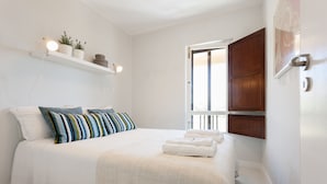 Un dormitorio con cama, almohadas, una mesita de noche, una estantería con plantas y una ventana con vistas.