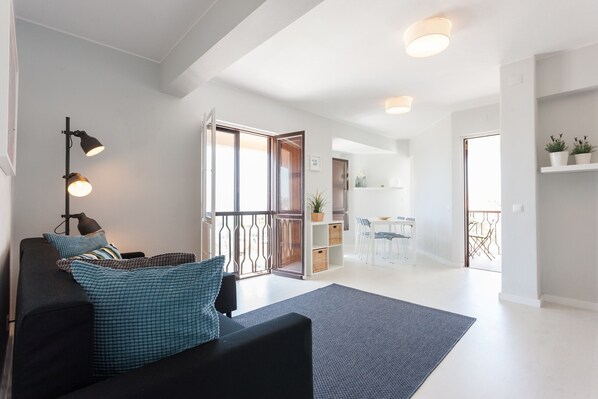 A modern living room with a black sofa, a blue rug, and a dining area in the background.