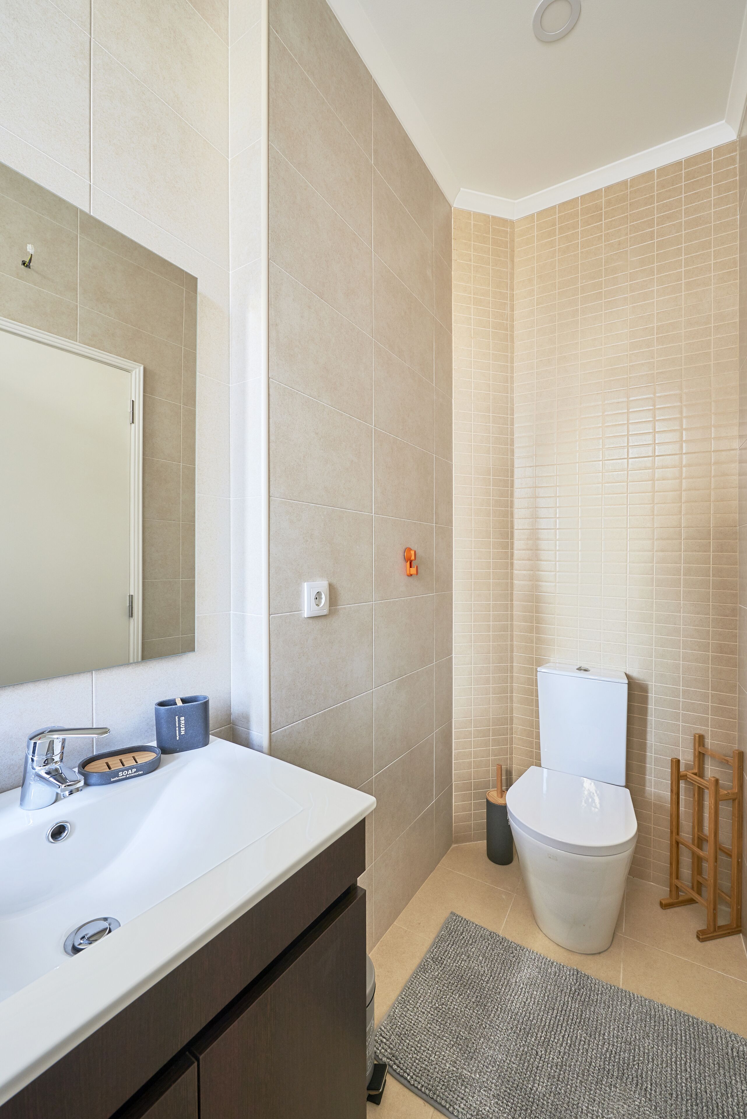 Un baño moderno con un lavamanos blanco, espejo y retrete, paredes con azulejos color beige y una escalera de madera.