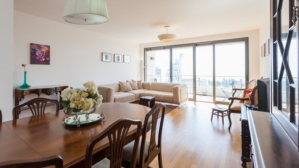 A spacious living room with a large dining table, a sofa, and a balcony with a view.