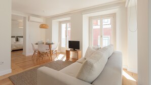 Een ruime woonkamer met een bank, een eettafel met stoelen, een televisie op een standaard en grote ramen voor natuurlijk licht.