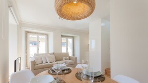 Een eetgedeelte met een ronde tafel gedekt voor vier personen, omringd door witte stoelen. Achtergrond is een beige bank, en de kamer heeft grote ramen.