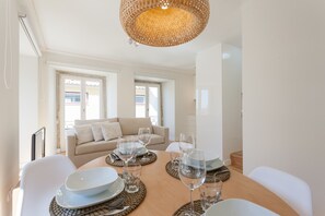 A dining area with a round table set for four, surrounded by white chairs. There is a beige sofa in the background, and the room has large windows.