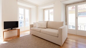 Een woonkamer met een beige bank, een flatscreen televisie op een houten standaard, en een groot raam met uitzicht op een balkon.