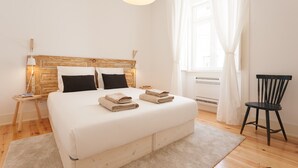 A bedroom with a wooden headboard, a bed with white linens, a bedside table with a lamp, a chair, and a window with curtains. - FLH Bica Modern Flat with View (Lisbon)
