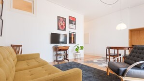 A living room with a yellow sofa, a black leather chair, a wooden side table, a flat-screen TV, and a potted plant. - FLH Bica Modern Flat with View (Lisbon)