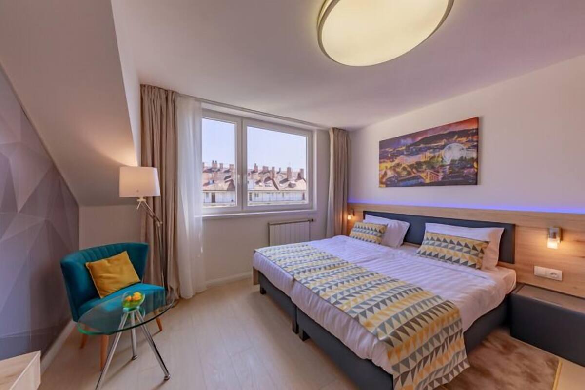 A hotel room with a bed, a chair, a table, a window with curtains, a painting on the wall, and a ceiling light.