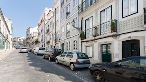 A narrow cobblestone street lined with multi-story buildings, parked cars, and balconies with potted plants. - FLH Príncipe Real Design Flat (Lisbon)