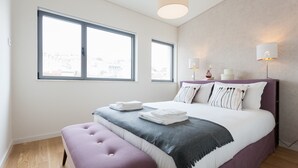 A modern bedroom with a large bed, two windows, a bedside table with lamps, and a view of the city.