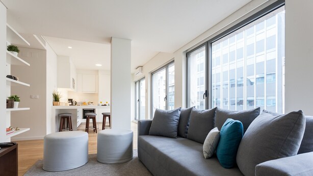 A modern living room with a grey sofa, ottomans, and a kitchen in the background.
