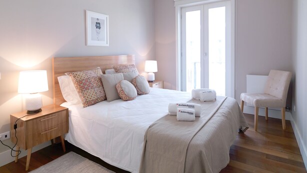 Een keurig opgemaakt slaapkamer met een bed, bedlampjes, nachtkastje, stoel en een raam met uitzicht.