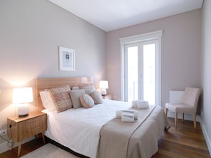 A neatly arranged bedroom with a bed, bedside lamps, a nightstand, a chair, and a window with a view.