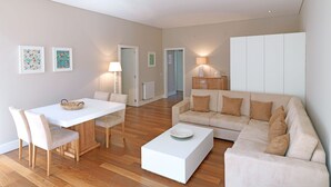Een moderne woonkamer met een witte bank, een eettafel met stoelen en een salontafel. Er hangen afbeeldingen in lijsten aan de muur, een lamp en een radiator.