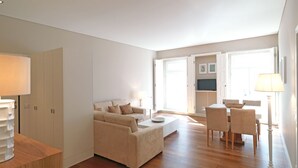Een moderne woonkamer met een bank, een eethoek met stoelen, een televisie en een groot raam.