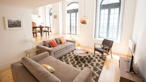 A modern living room with a sofa, armchair, and coffee table. There are large windows with arched tops, a dining area in the background, and a fireplace. - FLH Santa Catarina Modern Place (Lisboa ciudad)