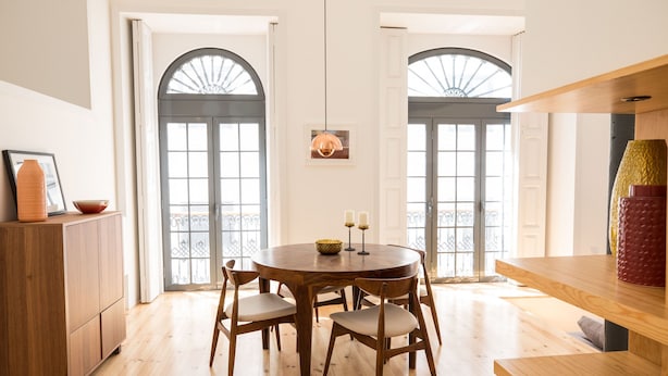 A dining area with a round wooden table, six chairs, and a cabinet. There are two large windows with arched tops and a hanging pendant light. - FLH Santa Catarina Modern Place (Lisboa ciudad)