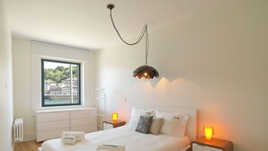 A bedroom with a large bed, two bedside tables with lamps, a radiator, a window with a view of buildings, and a hanging light fixture. - FLH Luxury Porto River View (Vila Nova de Gaia)