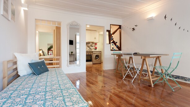 Un dormitorio con una cama, piso de madera, una zona de comedor con mesa y sillas, y una cocina al fondo.