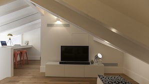 A modern loft apartment with a sloped ceiling, a flat-screen TV mounted on a white entertainment unit, a small desk with a laptop, and a cozy seating area with patterned cushions. - FLH Restauradores Luxus Flat (Lisbon)