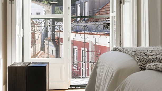 A bedroom with a bed, a small wooden cabinet, and a view of buildings through an open door. - FLH Restauradores Luxus Flat (Lisbon)