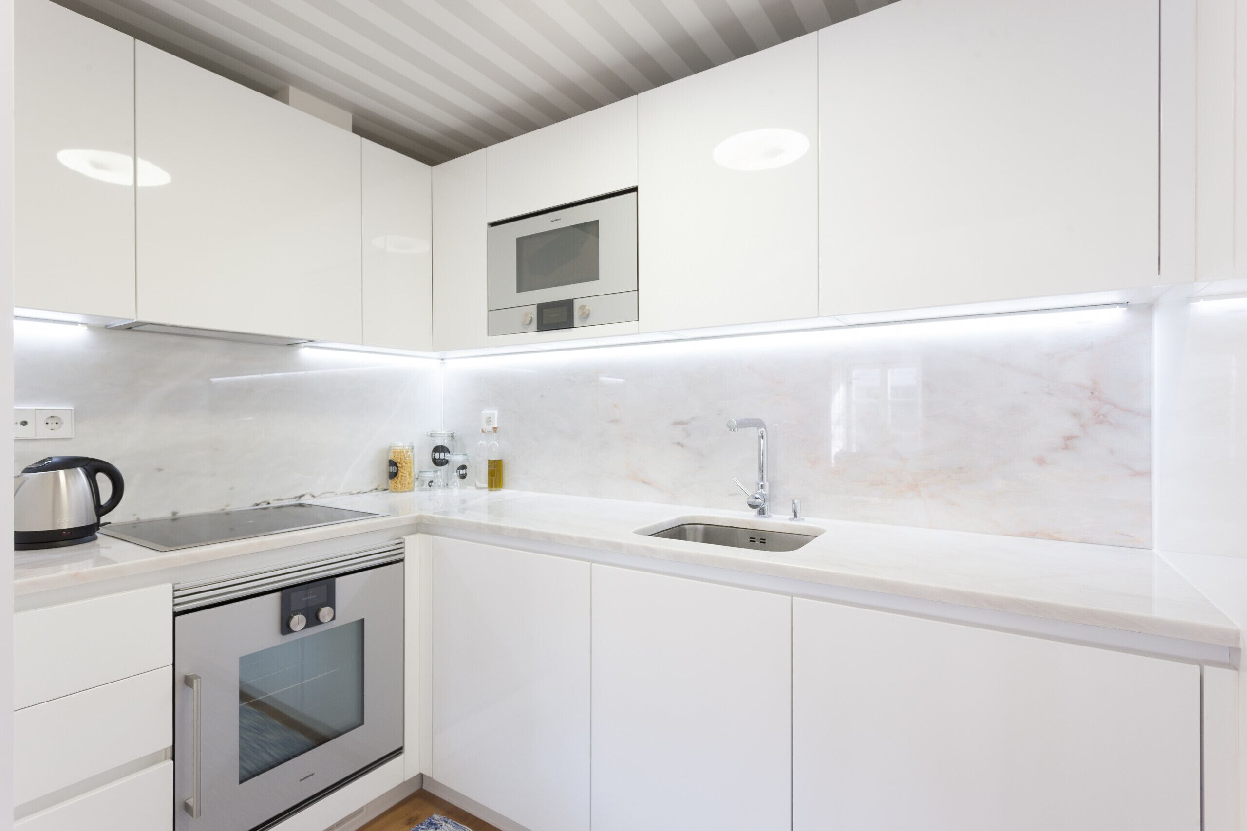 Une cuisine moderne avec des meubles blancs, un four encastré, un four à micro-ondes et un évier en acier inoxydable.