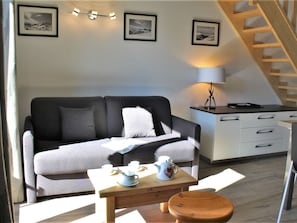 Uma sala de estar acolhedora com um sofá, uma mesa de centro e uma luminária. Há quadros nas paredes e uma escada ao fundo.
