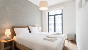 Ein modernes Hotelzimmer mit zwei Betten, einem großen Fenster, einem kleinen Hocker und einem Nachttisch mit Lampe.