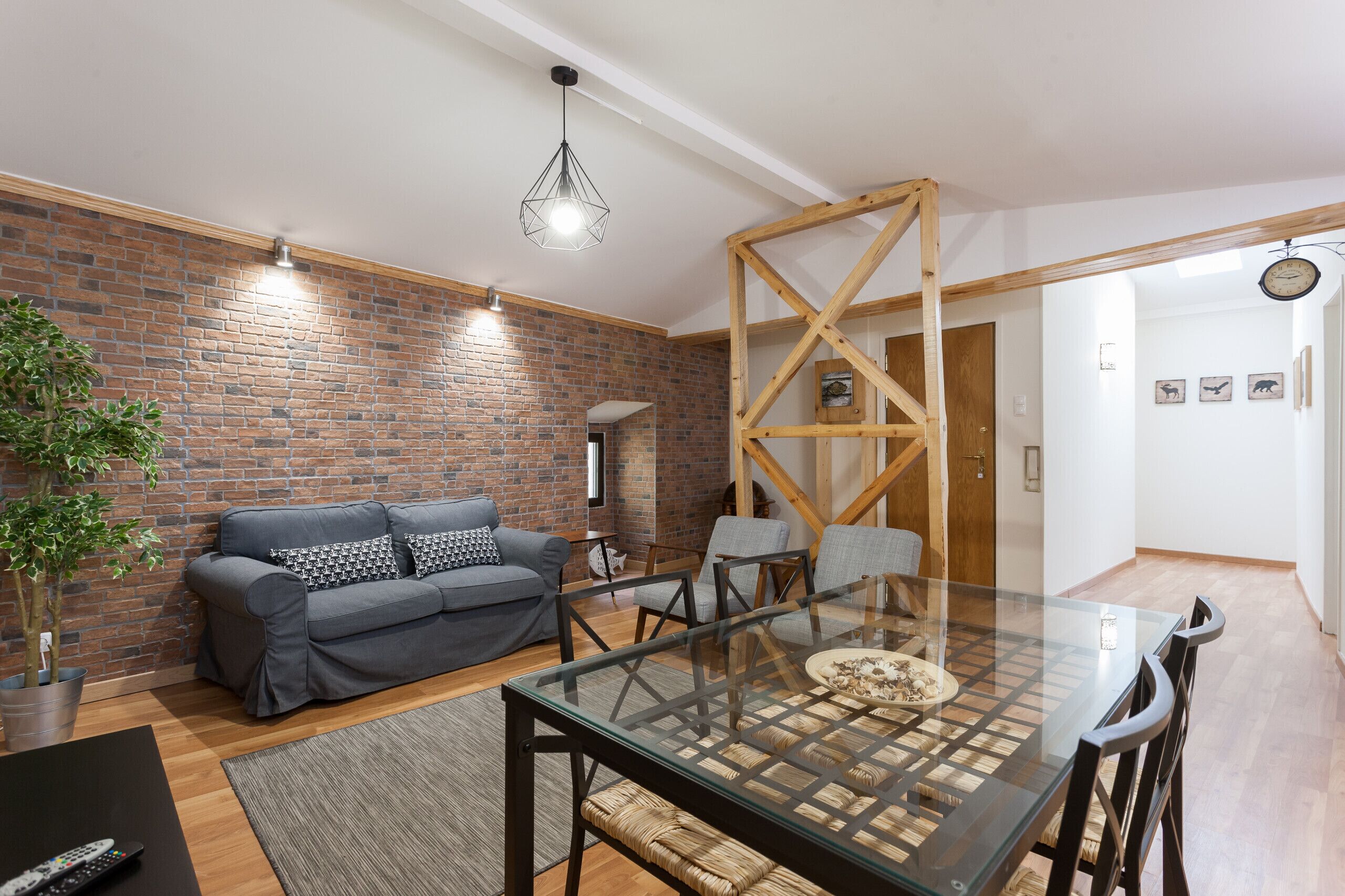 A modern living room with a brick wall, a gray sofa, a glass dining table, and a wooden ceiling structure.