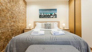 A neatly made bed with a patterned bedspread, a stone accent wall, a painting of a coastal scene, and a wooden wardrobe. - FLH Bairro Alto Stylish Apartment (Lisbon)