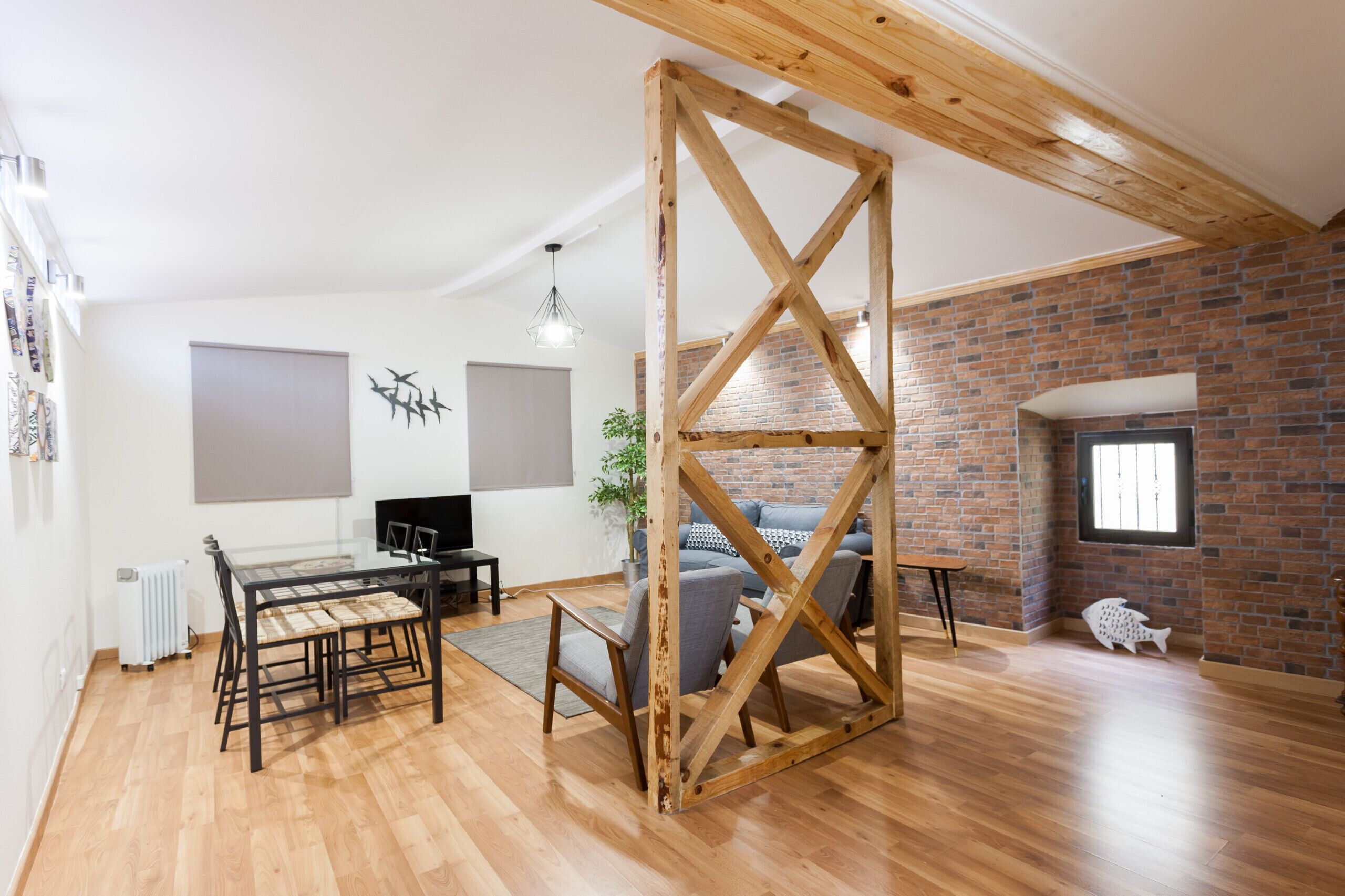 Ein geräumiger Raum mit sichtbarer Holzdecke, einem Essbereich mit Tisch und Stühlen, einem grauen Sofa und einer Backsteinwand.