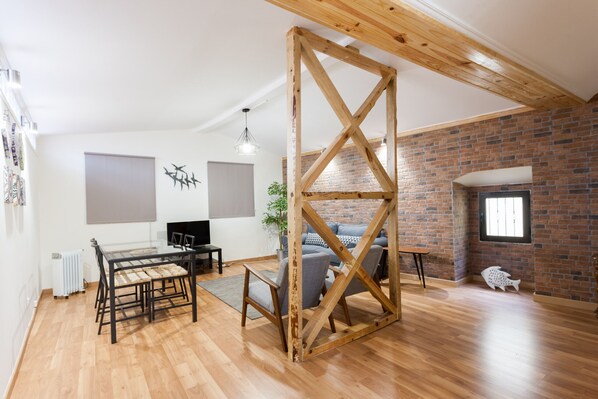 A spacious room with a wooden ceiling structure, a dining area with a table and chairs, a gray sofa, and a brick wall.