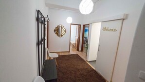 Un pasillo con una alfombra marrón, una silla blanca, una mesa negra, un perchero metálico, un espejo redondo y un cartel que dice 'Welcome'.