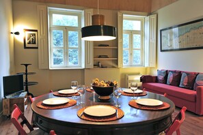 A dining area with a round table set for four, surrounded by red chairs. A large bowl of fruit is placed in the center of the table.