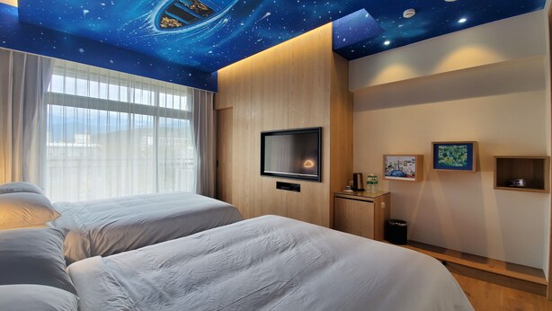 Habitación de hotel con diseño de techo estrellado, dos camas, un televisor y una mesita con un marco de fotos y un cesto de basura.