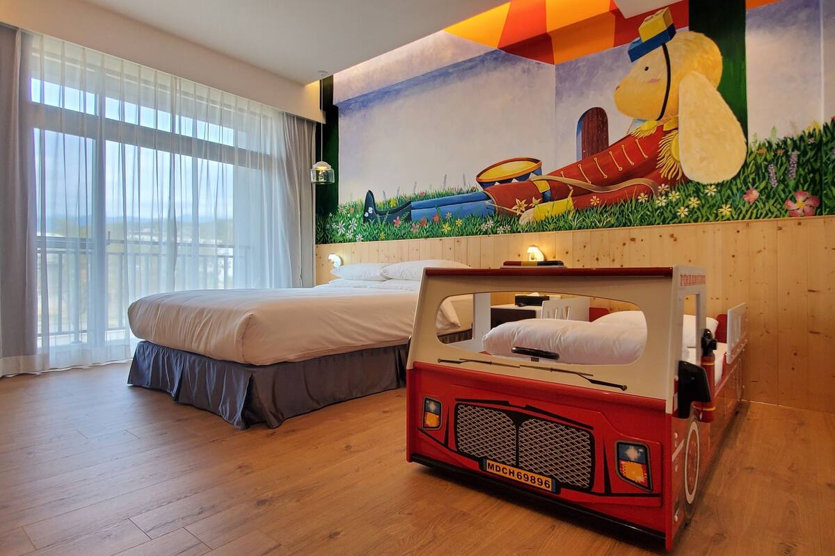 A bedroom with a bed, a unique red vehicle-shaped bed, a large window with sheer curtains, and a vibrant mural on the wall.