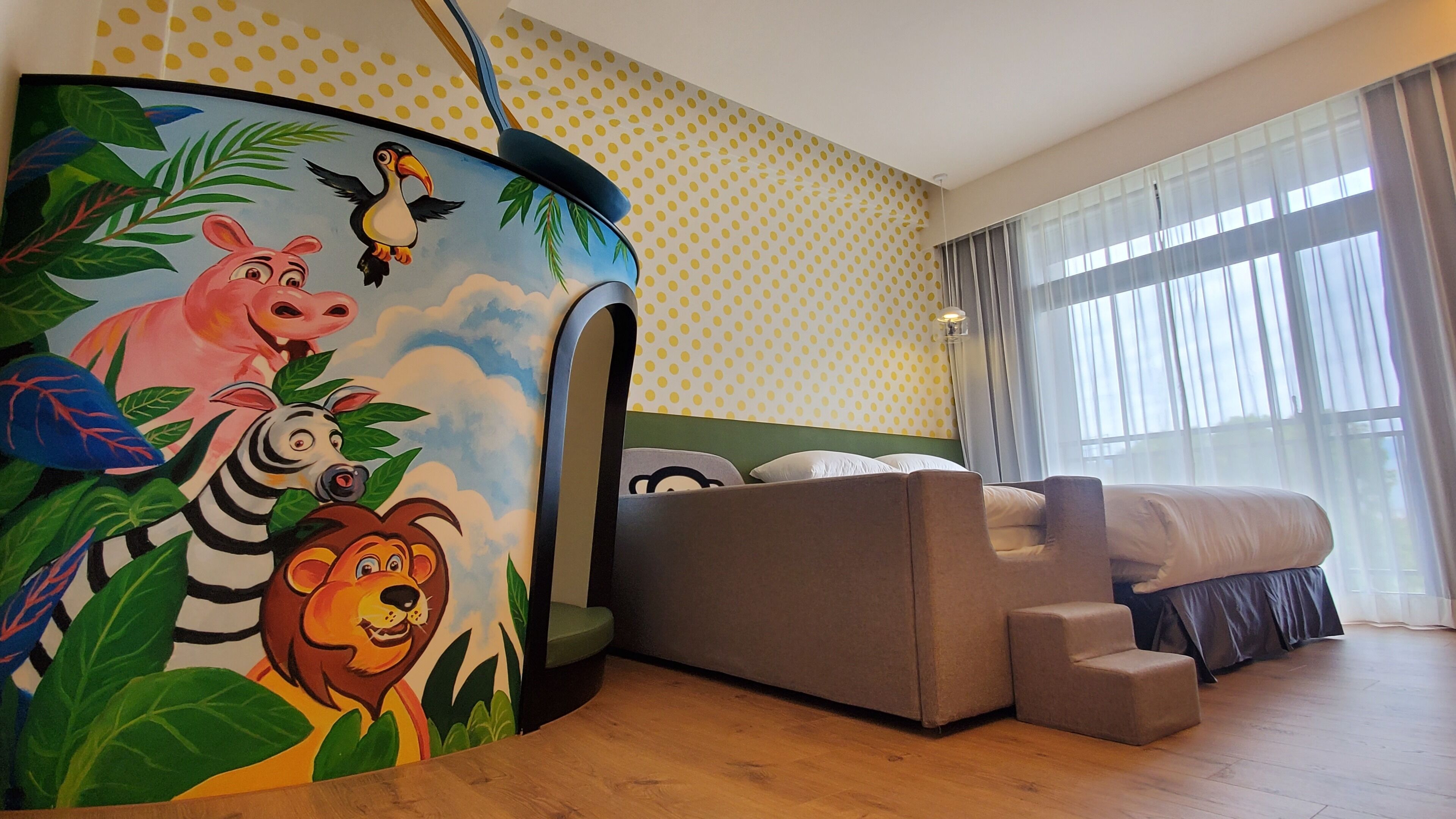 Quarto de hotel com cama, sofá, parede com padrão de bolinhas e mural com ilustrações de animais.