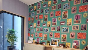 Un salón moderno con una pared verde decorada con obras de arte coloridas, un ventanal grande y un área de estar con un sofá y dos mesas redondas.
