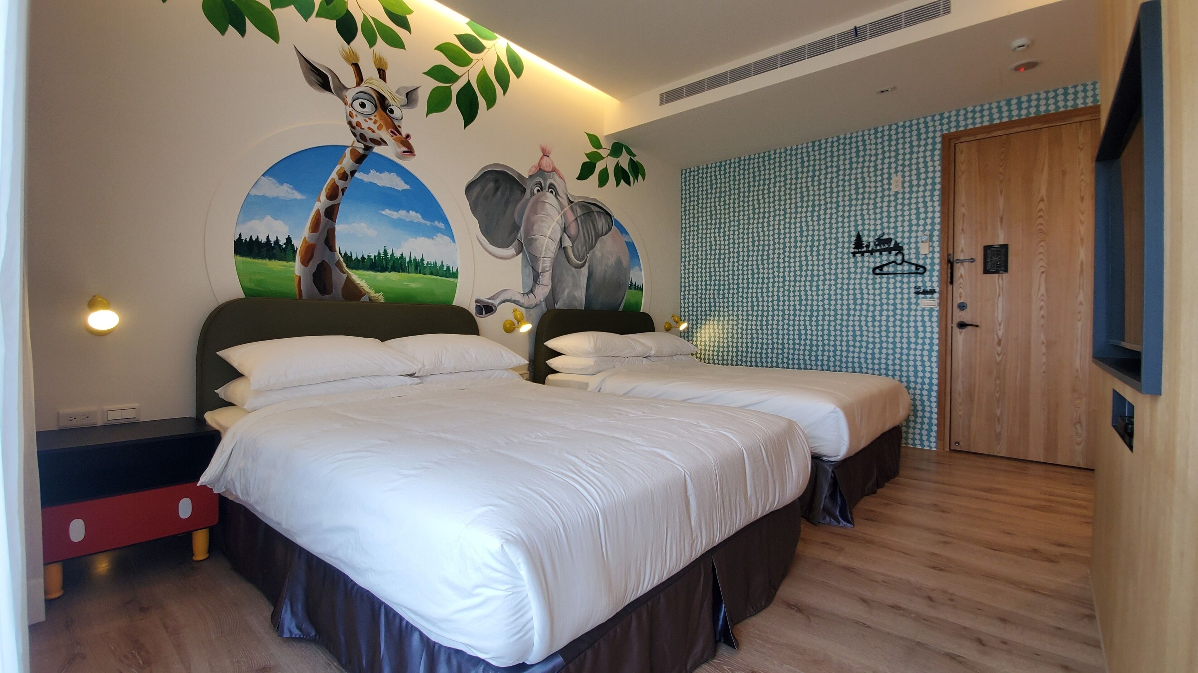 Une chambre d’hôtel avec deux lits, une fresque représentant une girafe et un éléphant, une porte en bois et un mur orné d’un motif.