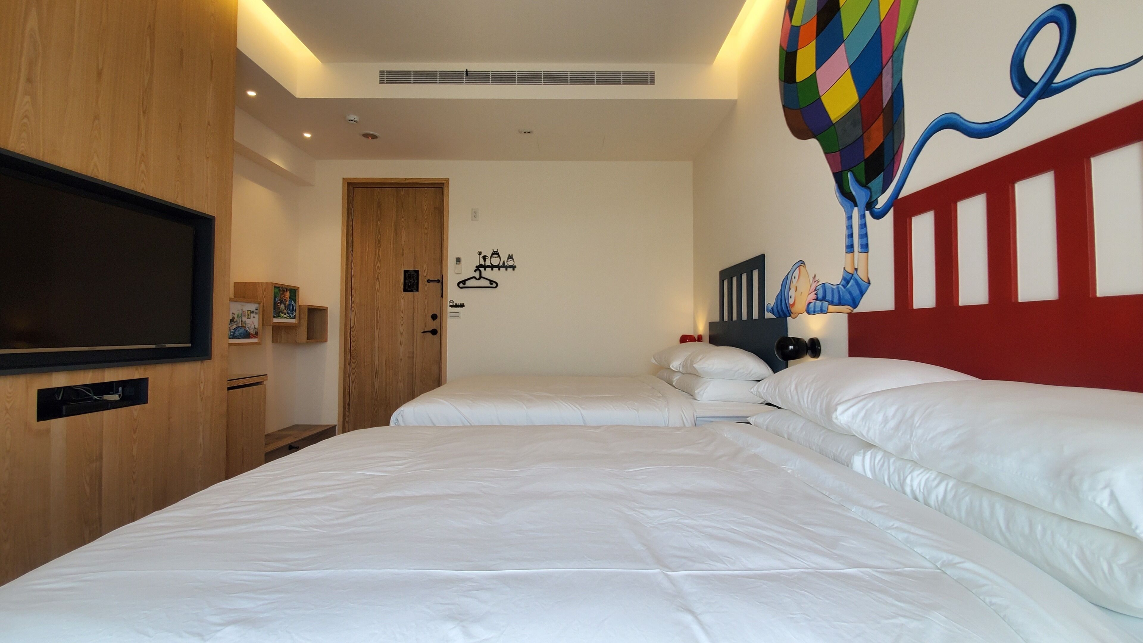 Een hotelkamer met twee bedden, een televisie, een houten kledingkast en een kleurrijke wanddecoratie.