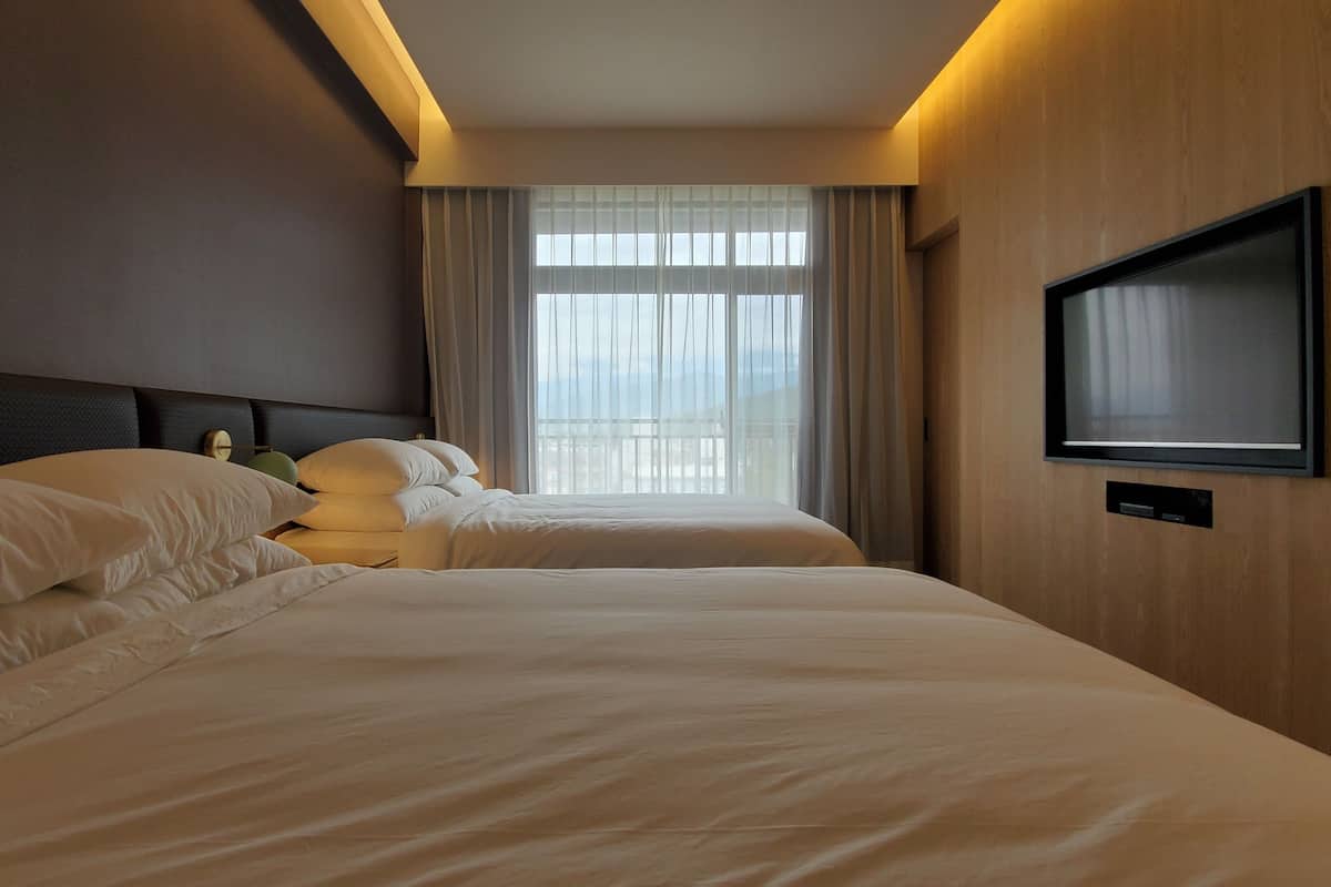 A hotel room with two beds, a flat-screen TV mounted on the wall, and a large window with curtains.