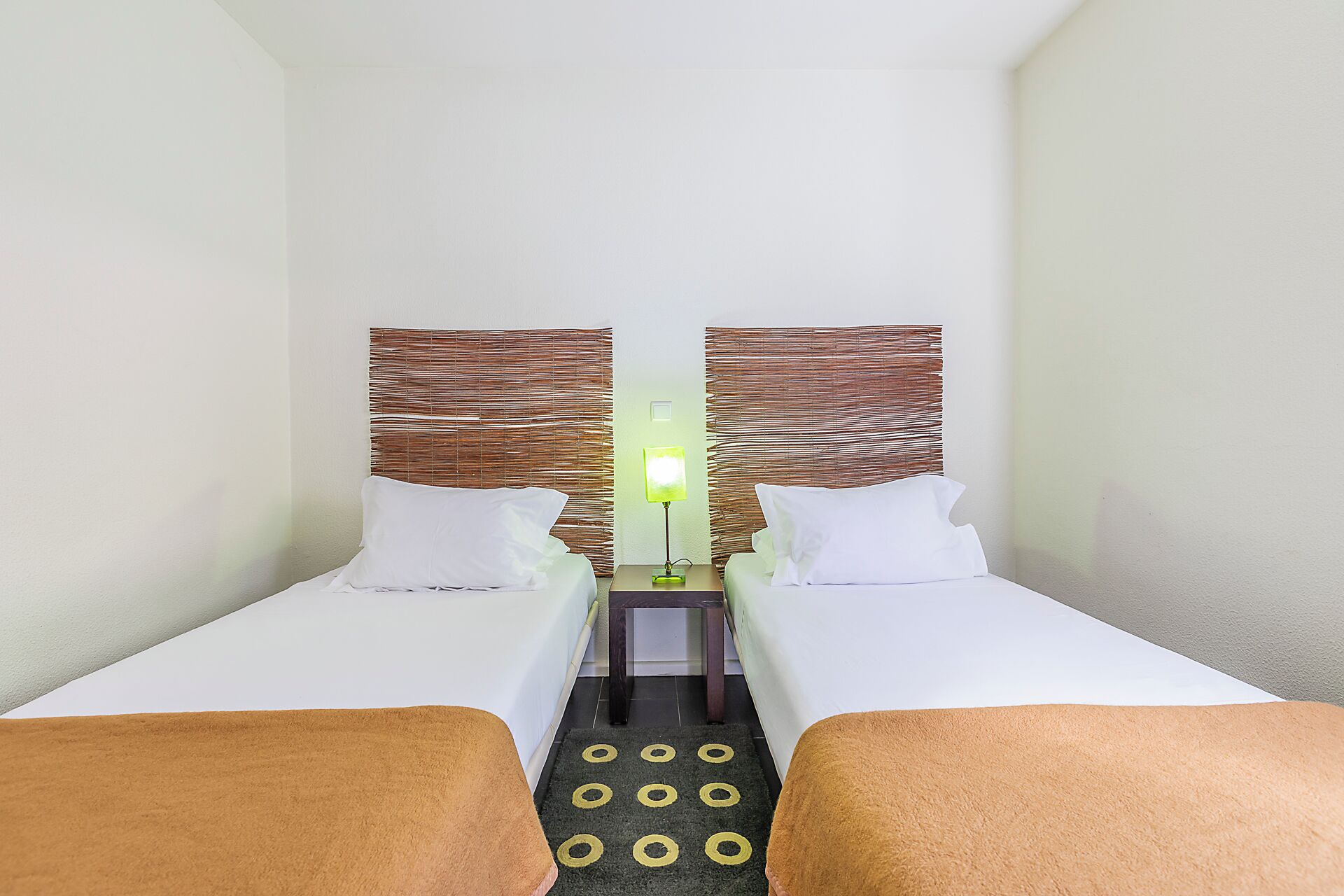Quarto de hotel com duas camas, uma mesa de cabeceira, um abajur e um tapete pequeno com padrões circulares.