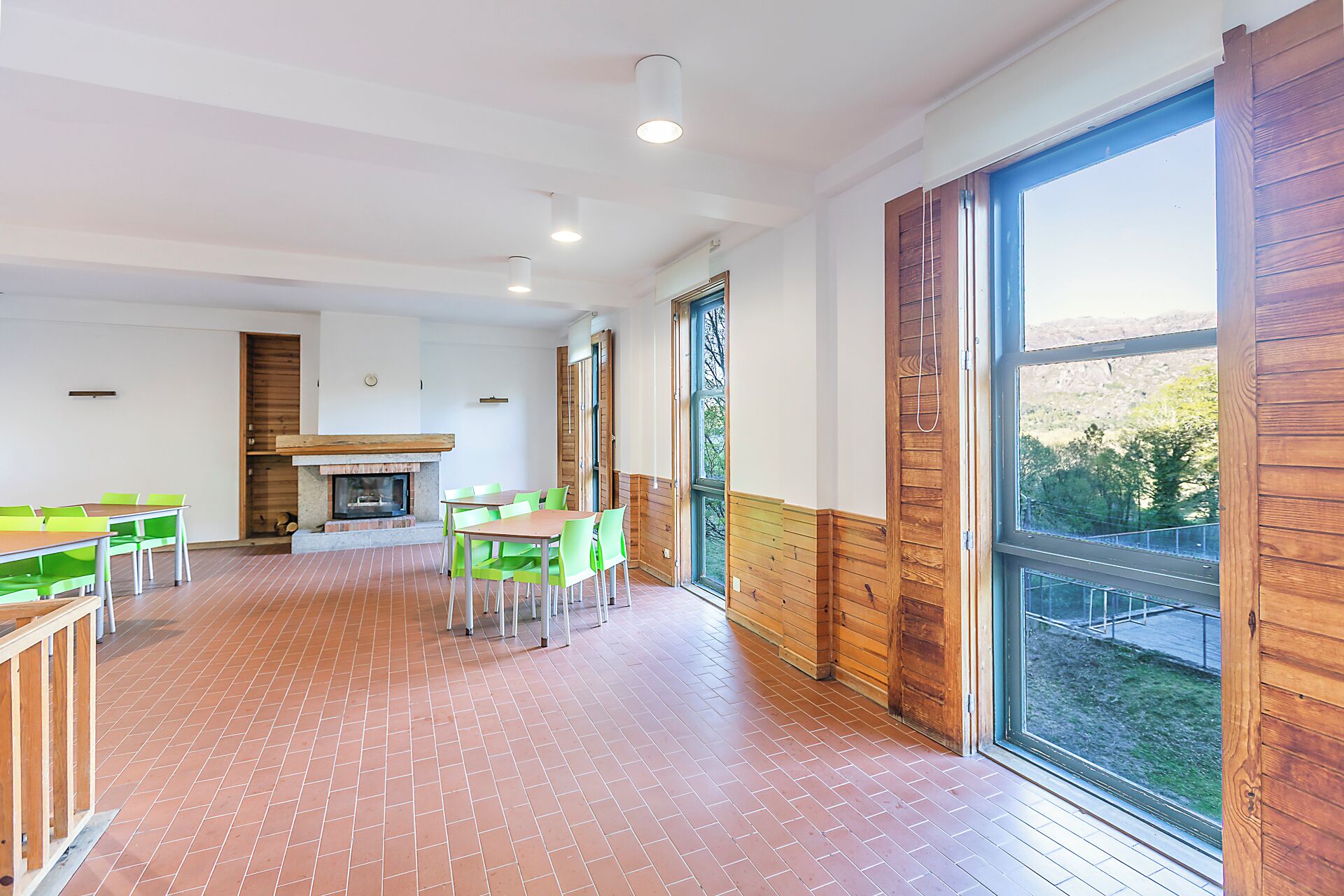 A spacious room with a fireplace, green chairs, a wooden table, and large windows.
